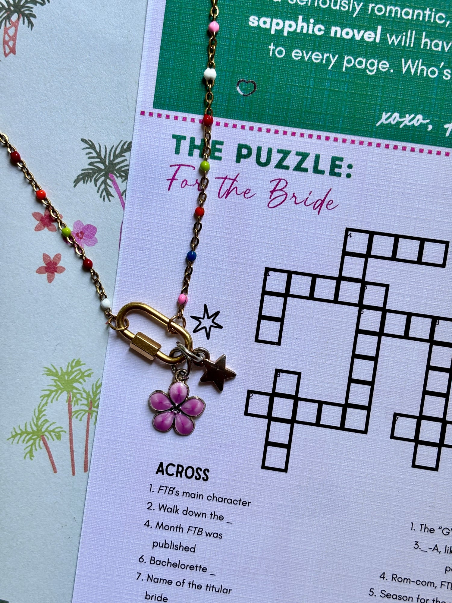 A close-up of a jewelry puzzle keychain with a gold-toned carabiner lock, a pink flower charm, and a star charm, laid over a printed crossword puzzle sheet titled 'THE PUZZLE: For the Bride'.
